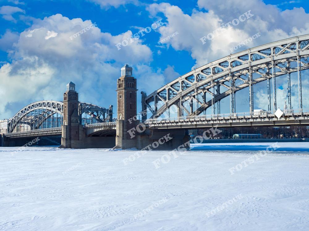 На фотографии — Большеохтинский мост, один из старейших разводных мостов Санкт-Петербурга. Мост соединяет центральную часть города с Малой Охтой и впечатляет массивными башнями и металлическими конструкциями. Под мостом — замёрзшая Нева, укрытая снежным покровом. Небо над городом ясное, с живописными облаками, что придаёт пейзажу яркость и глубину. Это символ технического прогресса и архитектурной красоты Северной столицы.